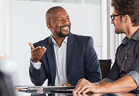 Two happy business colleagues at meeting in modern office interior. Successful african boss in a conversation with young employee in boardroom. Marketing team of two businessmen discussing strategy.